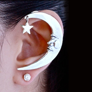 May include: A silver crescent moon ear cuff with a dangling star charm. The moon has a face sculpted on it. A small, clear, round gemstone stud is in the earlobe.
