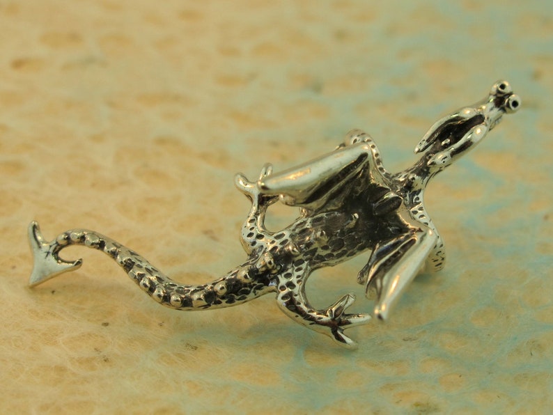 May include: A detailed silver dragon figurine with outstretched wings and a long tail. The dragon has textured scales and is posed as if in flight. The figurine is set against a light-colored background.