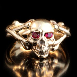 May include: A gold skull ring with two ruby red eyes and a crossbones design.