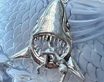 May include: Silver-toned shark pendant necklace. The shark's mouth is open, revealing sharp teeth and a small figure. The pendant hangs from a silver chain. The background is a textured, light blue surface.