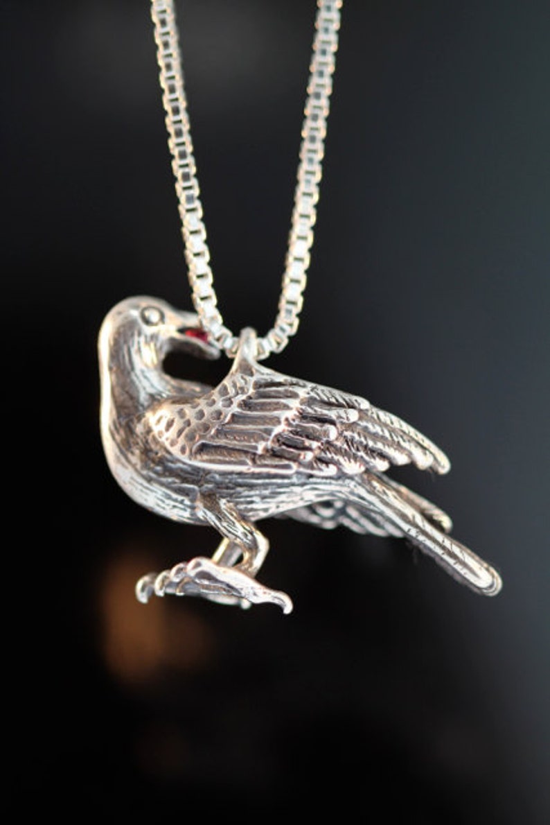 May include: A silver pendant necklace featuring a detailed, three-dimensional crow with a red gemstone in its beak.