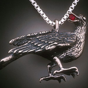 May include: A silver crow pendant with a red gemstone in its beak. The crow is perched with its wings spread and its feet outstretched.