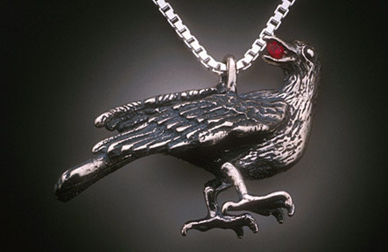 May include: A silver crow pendant with a red gemstone in its beak. The crow is perched with its wings spread and its feet outstretched.
