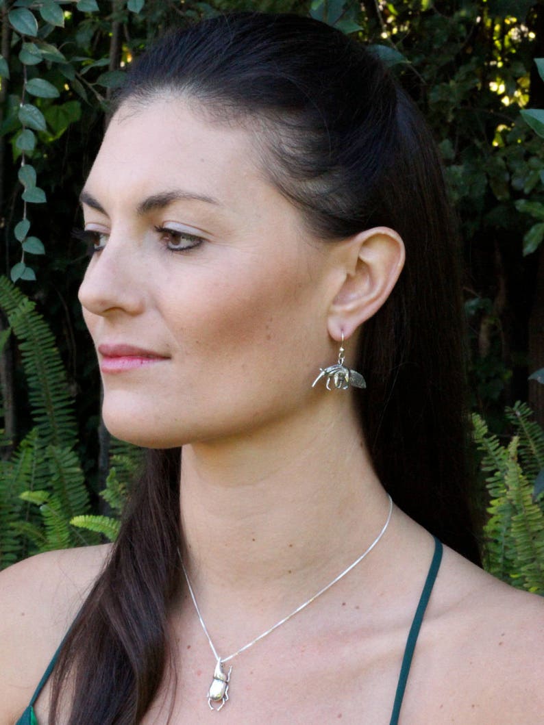 May include: A woman wearing a green tank top and a silver necklace with a beetle pendant. She is also wearing silver earrings with a beetle design.
