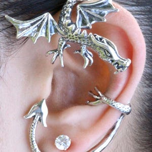 May include: A silver dragon ear cuff with detailed features. The dragon has outstretched wings, a detailed head, and a tail that curves around the ear. A small, clear stud earring is also visible.