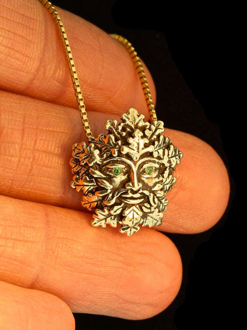 Op de afbeelding: Een gouden ketting met een groene man hanger. De hanger is gemaakt van metaal en heeft een gezicht met groene ogen en een baard van bladeren. De ketting is aan een ketting met een vierkant schakels patroon.