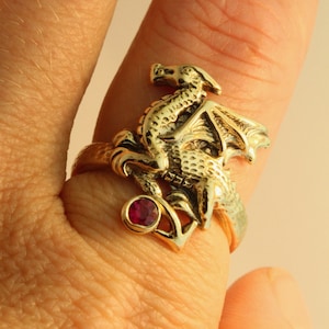 Puede incluir: Un anillo de oro con un diseño de dragón detallado y una piedra preciosa roja. El dragón está posado en una rama con las alas extendidas.