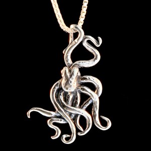 May include: A silver octopus pendant with a textured surface hangs from a silver chain. The octopus is curled up with its tentacles wrapped around itself.