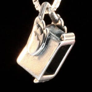 May include: A silver charm necklace pendant shaped like a small, open-top box with a hinged lid. The box has a detailed design and a smooth, polished finish.