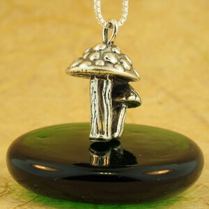 May include: A silver mushroom charm pendant with a detailed textured stem and a domed cap. The pendant is hanging from a silver chain and is resting on a green glass surface.
