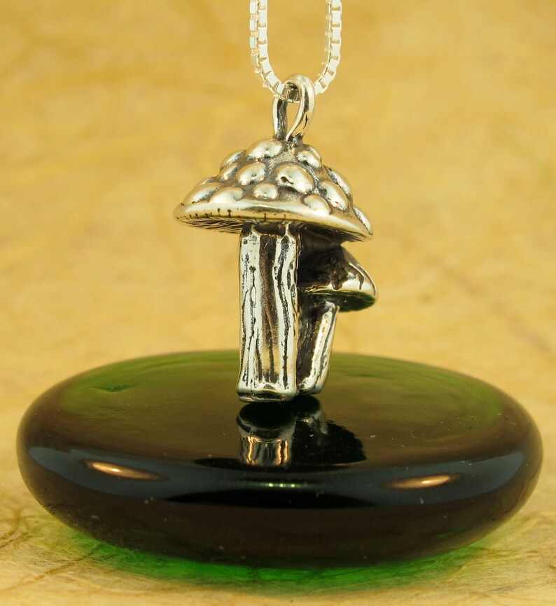 May include: A silver mushroom charm pendant with a detailed textured stem and a domed cap. The pendant is hanging from a silver chain and is resting on a green glass surface.