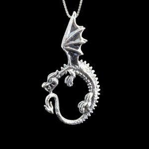 Op de afbeelding: Zilveren ketting met hanger met een gedetailleerd drakenontwerp met uitgespreide vleugels en een staart die om zijn lichaam is gekruld.