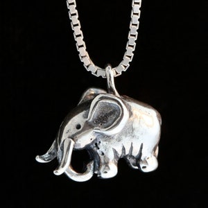 Puede incluir: Un collar de plata con un dije de elefante y una cadena de eslabones cuadrados. El dije de elefante está detallado con una trompa, orejas y pies.