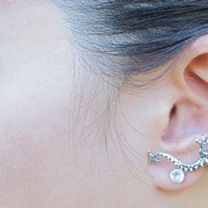 May include: A silver-toned ear cuff featuring a detailed dragon design. The dragon wraps around the ear, with a clear, round gemstone accent. The jewelry is worn on the ear, showcasing the intricate craftsmanship of the dragon's scales and form.