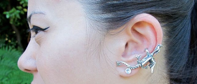 May include: A silver-toned ear cuff featuring a detailed dragon design. The dragon wraps around the ear, with a clear, round gemstone accent. The jewelry is worn on the ear, showcasing the intricate craftsmanship of the dragon's scales and form.