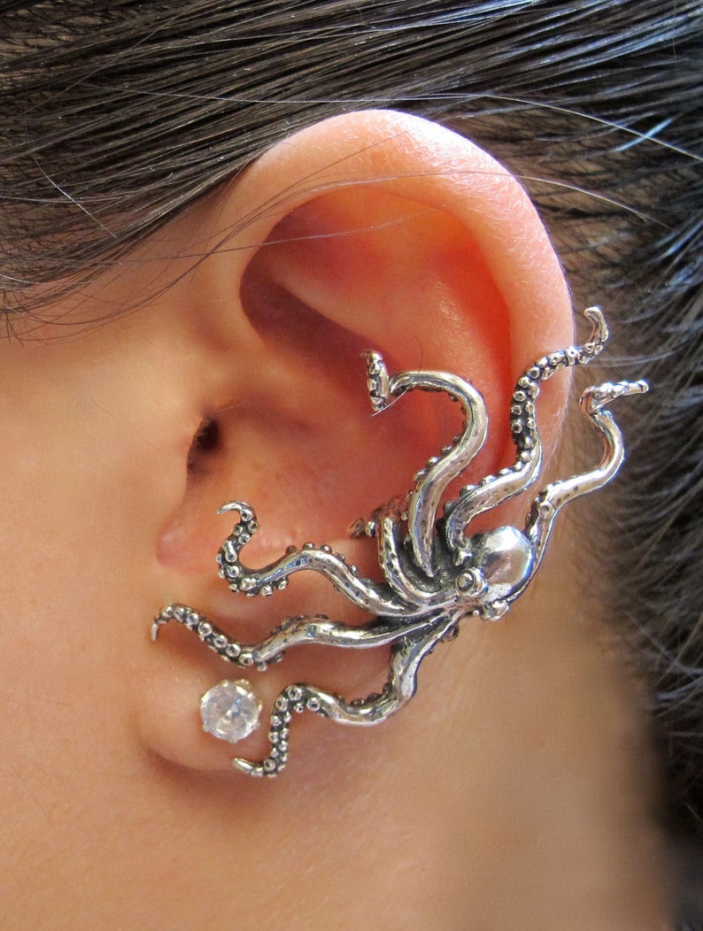 Puede incluir: Un pendiente de oreja de plata en forma de pulpo con un pendiente de cristal transparente. El pulpo tiene un dise&ntilde;o detallado con tent&aacute;culos que se envuelven alrededor de la oreja.