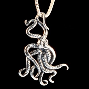 May include: A silver octopus pendant with a textured surface hangs from a silver chain. The pendant is detailed with eight tentacles.