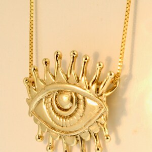 May include: A gold chain necklace with a pendant featuring a stylized eye with a crown of rays. The eye is detailed with a pupil and iris.