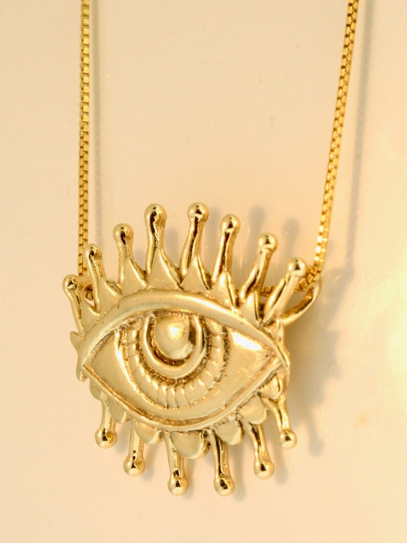 May include: A gold chain necklace with a pendant featuring a stylized eye with a crown of rays. The eye is detailed with a pupil and iris.