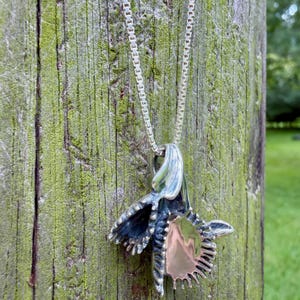 K&ouml;nnte beinhalten: Ein silberner Anh&auml;nger in Form einer stilisierten Orchideenbl&uuml;te mit polierter Metalloberfl&auml;che und dunklen, oxidierten Vertiefungen, der an einer zarten Silberkette mit quadratischen Gliedern h&auml;ngt, die eine glatte und reflektierende Oberfl&auml;che aufweist.