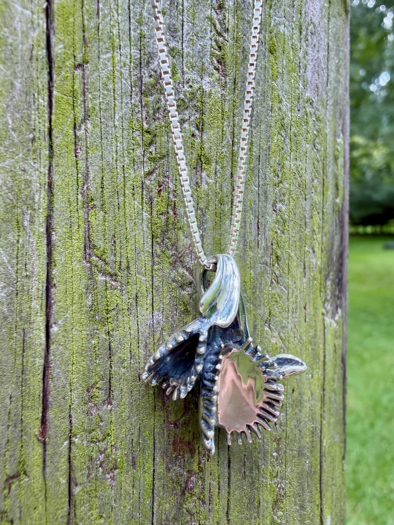 K&ouml;nnte beinhalten: Ein silberner Anh&auml;nger in Form einer stilisierten Orchideenbl&uuml;te mit polierter Metalloberfl&auml;che und dunklen, oxidierten Vertiefungen, der an einer zarten Silberkette mit quadratischen Gliedern h&auml;ngt, die eine glatte und reflektierende Oberfl&auml;che aufweist.
