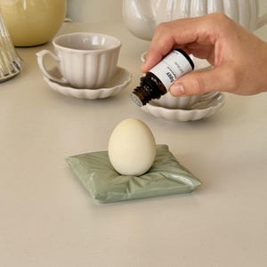 May include: A white egg sits on a small, sage green pillow-shaped ceramic dish. A hand holds a small brown bottle with a white label, pouring liquid onto the egg. Teacups and other white ceramic items are in the background.