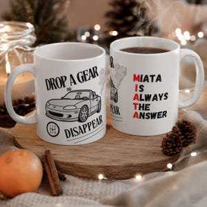 May include: Two white ceramic mugs featuring a black line drawing of a convertible car. The text reads "DROP A GEAR AND DISAPPEAR" with a gear shift pattern icon, and "MIATA IS ALWAYS THE ANSWER" with the word MIATA highlighted in red letters.