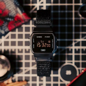 May include: A black Casio F-91W digital watch with a black fabric strap. The watch face displays the time as 15:32:00, with the date of May 18. The watch is set on a gray and white grid surface.