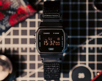 Könnte beinhalten: Eine schwarze digitale Casio F-91W-Uhr mit einem schwarzen Stoffarmband. Das Zifferblatt zeigt die Uhrzeit 15:32:00 mit dem Datum 18. Mai an. Die Uhr liegt auf einer grau-weißen Rasteroberfläche.