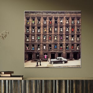 May include: A black and white vintage photograph of a city street with a black car parked in front of a building. The building has many windows with people looking out of them. The people are all wearing different coloured clothing.