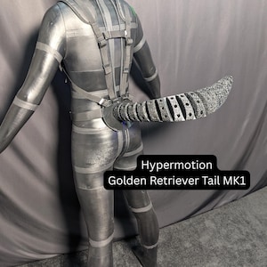 May include: A black articulated robotic tail attached to a harness worn on a mannequin. The tail features a segmented, textured design with visible circular joints. A black label with white text reads: Hypermotion Golden Retriever Tail MK1.