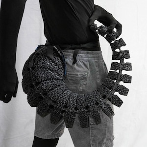 May include: A black, segmented, sculptural accessory, possibly a belt or harness, with a textured, woven appearance. The piece is worn around the waist and extends outward, creating a unique, avant-garde silhouette. The design features a series of interconnected, rectangular elements.