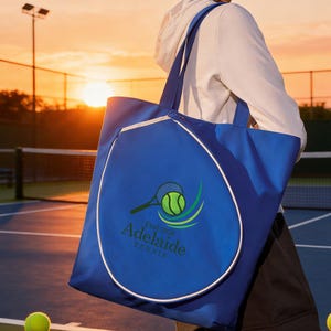 Könnte beinhalten: Eine blaue Tennis-Tragetasche mit weißer Paspelierung und Griffen. Die Tasche zeigt ein Tennis-Schläger- und Ball-Logo mit dem Text "Adelaide Tennis Est. 2026". Die Tasche wird auf einem Tennisplatz getragen.