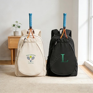 May include: Two tennis backpacks, one cream-colored and one black, each with a tennis racket attached. The cream backpack has a tennis club logo and the text "The Lauren Tennis Club". The black backpack has a green "L" on it.