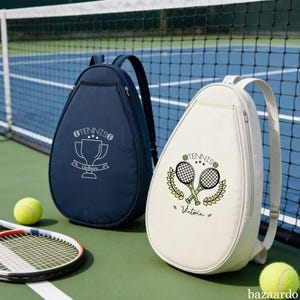 Könnte beinhalten: Zwei Tennisschlägertaschen, eine marineblaue und eine cremefarbene, mit Reißverschlüssen und verstellbaren Trägern. Die marineblaue Tasche hat eine Trophik-Grafik und das Wort "Tennis" aufgedruckt. Die cremefarbene Tasche hat gekreuzte Schläger und den Namen "Victoria".