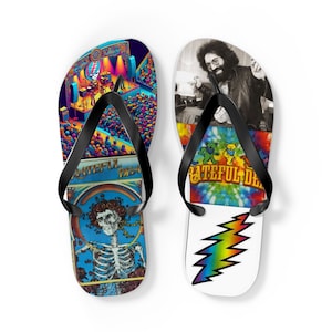 May include: Pair of black flip flops with Grateful Dead themed designs. One flip flop features a colorful illustration of a concert stage with a crowd of people, a skeleton with roses, and the text "Grateful Dead". The other flip flop features a tie-dye pattern, a dancing bear, and the text "Grateful Dead".