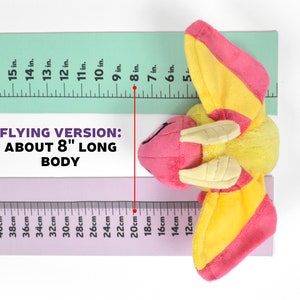 May include: A plush toy in the shape of a flying insect, primarily yellow and pink, with a body approximately 8 inches long. The toy is positioned next to a ruler for size comparison, with the text "FLYING VERSION: ABOUT 8" LONG BODY".