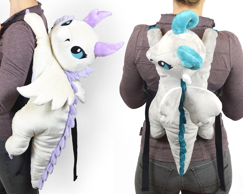 May include: Two plush dragon backpacks, one white with purple accents and one gray with teal accents. Both dragons have wings and tails and are designed to be worn on the back.