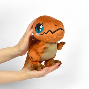 Puede incluir: Un peque&ntilde;o dinosaurio de peluche naranja con grandes ojos azules y una expresi&oacute;n linda. El dinosaurio est&aacute; de pie sobre sus dos patas traseras y tiene los brazos extendidos.