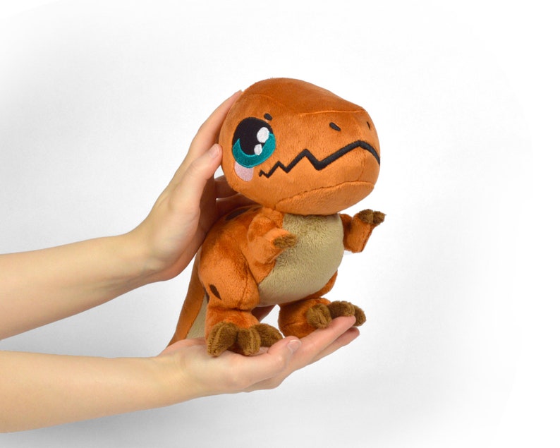 Puede incluir: Un peque&ntilde;o dinosaurio de peluche naranja con grandes ojos azules y una expresi&oacute;n linda. El dinosaurio est&aacute; de pie sobre sus dos patas traseras y tiene los brazos extendidos.