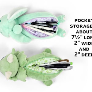 May include: Two plush pencil cases, one shaped like a dinosaur and the other like a bunny, both with open mouths revealing green fabric interiors. The dinosaur case has a green and white leaf pattern, while the bunny case has a purple and white floral pattern. Both cases are filled with pens and pencils. The text "POCKET STORAGE IS ABOUT 7 1/2" LONG 2" WIDE AND 2" DEEP" is visible in the image.