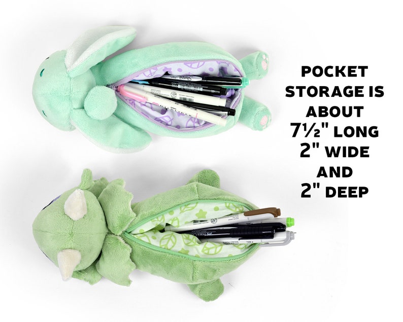 May include: Two plush pencil cases, one shaped like a dinosaur and the other like a bunny, both with open mouths revealing green fabric interiors. The dinosaur case has a green and white leaf pattern, while the bunny case has a purple and white floral pattern. Both cases are filled with pens and pencils. The text "POCKET STORAGE IS ABOUT 7 1/2" LONG 2" WIDE AND 2" DEEP" is visible in the image.