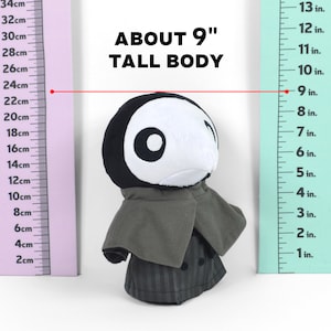 Puede incluir: Un juguete de peluche negro y blanco con un abrigo gris. El juguete mide aproximadamente 9 pulgadas de alto y est&aacute; de pie junto a una regla. La regla muestra que el juguete mide aproximadamente 23 cent&iacute;metros de alto.