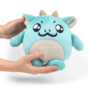Peut inclure: Un jouet en peluche de dragon bleu clair avec un visage souriant, de grands yeux et un petit ventre beige. Le dragon a de petites cornes beiges et est assis sur ses pattes arri&egrave;re.