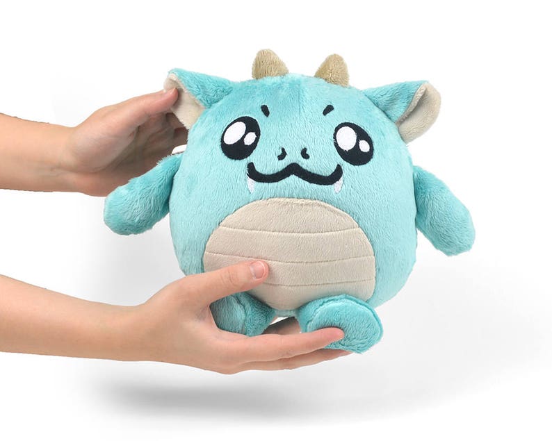 Peut inclure: Un jouet en peluche de dragon bleu clair avec un visage souriant, de grands yeux et un petit ventre beige. Le dragon a de petites cornes beiges et est assis sur ses pattes arri&egrave;re.