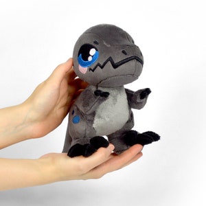 Puede incluir: Un peque&ntilde;o dinosaurio de peluche gris con ojos azules y nariz rosa. El dinosaurio est&aacute; de pie sobre sus patas traseras con las patas delanteras levantadas.