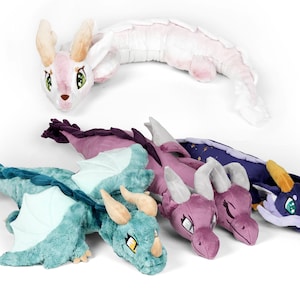 May include: Five plush dragon toys in different colors and designs. The dragons are all lying down with their wings spread out. The colors include pink, purple, teal, and blue.