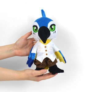 May include: A blue and yellow plush bird wearing a white shirt and brown pants. The bird has a black beak and green eyes.