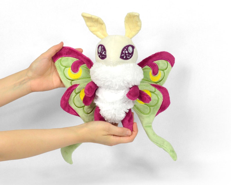 May include: A plush Luna moth toy with a cream-colored body, large green and magenta wings, and yellow accents. The moth has large, detailed eyes and a fluffy white chest. The toy is held by two hands.