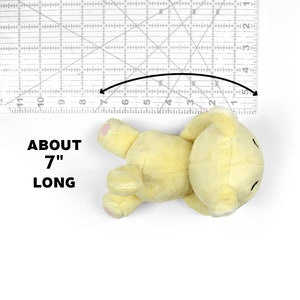 May include: A yellow plush teddy bear lying on its back, measuring approximately 7 inches long. The bear has a simple face with black eyes and a small mouth.